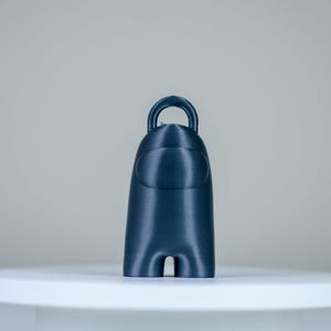 May include: A navy blue figurine resembling a character from the game Among Us. The figure has a rounded body, a visor-like face, and a loop at the top. It stands on a white surface against a neutral background.