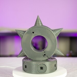 May include: A gray, star-shaped object with conical spikes and a central circular opening. The object is mounted on a cylindrical base with square cutouts. The background is a blurred purple.