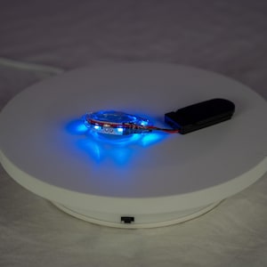 May include: A circular white rotating display stand featuring a small, illuminated blue LED light component connected by thin red and black wires to a rectangular black battery pack resting on the surface of the stand.