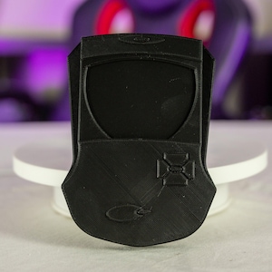 May include: A black 3D-printed phone case with a unique design. The case features a shield-like shape with a cross-shaped design and a curved emblem. The case is displayed on a white circular stand.