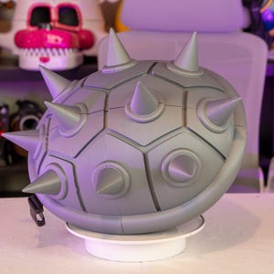 May include: A gray, spiky Koopa Troopa shell prop, a video game accessory. The shell has a segmented design with rounded edges and conical spikes. It is displayed on a white rotating stand, with other collectibles in the background.