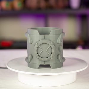 Puede incluir: Un recipiente cilíndrico gris con un emblema circular con un símbolo 'X'. El recipiente tiene elementos decorativos redondeados y se muestra sobre una plataforma giratoria blanca. El artículo parece estar impreso en 3D.