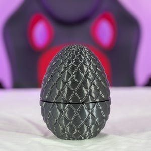 May include: A dark gray, dragon egg-shaped container with a textured, scale-like pattern. The egg is split in two, revealing a possible storage compartment. The background is blurred, with a purple and black color scheme.