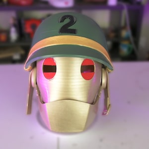 May include: A gold-colored, robotic-style helmet with red eye openings. The helmet has a green top with the number "2" in black and a gold band. The faceplate is gold, and the design suggests a futuristic or cosplay theme.