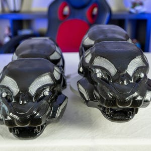 Puede incluir: Cuatro esculturas decorativas negras y plateadas de una cabeza de animal estilizada con bocas abiertas y detalles. Las esculturas están dispuestas sobre una superficie blanca, con un fondo borroso.