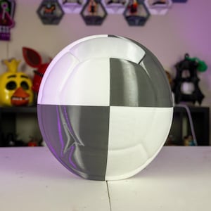 May include: A spherical object divided into four equal sections, alternating between white and gray. The object is smooth and reflective, with a slight sheen. The background includes decorative items.
