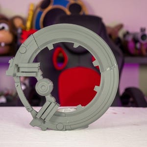 May include: A gray, circular, 3D-printed object with a segmented design. The object has a central circular opening and several rectangular and circular components attached to its frame. The object is resting on a white surface.