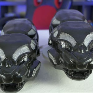 May include: Four black panther head sculptures with silver accents are displayed on a white surface. The sculptures are detailed, with visible eyes, noses, and open mouths. The heads are arranged in two pairs, suggesting a set or collection.