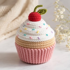 May include: A crocheted cupcake with a red cherry on top. The cupcake has white frosting with colorful sprinkles, a tan cake base, and a pink cupcake liner. The cherry has green leaves.