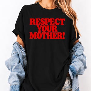 Può includere: Una t-shirt nera a maniche corte con la scritta RESPECT YOUR MOTHER! stampata in grassetto rosso a lettere maiuscole sul petto. La maglietta ha uno scollo rotondo ed è indossata con una giacca di jeans azzurra sulle spalle.