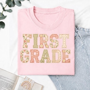 Può includere: Una t-shirt rosa chiaro con il testo FIRST GRADE in grandi lettere stampatello. Le lettere presentano un design patchwork con vari motivi, tra cui stampe floreali, quadretti vichy e pois in tonalità rosa, verde e crema.