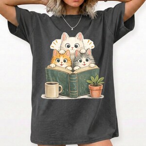 Può includere: Una t-shirt a maniche corte grigio antracite con un'illustrazione grafica di tre gattini, uno bianco, uno arancione e uno tigrato grigio, che sbucano da un libro verde aperto. Una tazza beige e una piccola pianta in vaso sono accanto al libro.