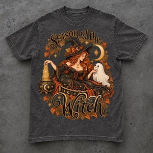 May include: A charcoal gray t-shirt featuring a graphic of a witch with long red hair wearing a hat, sitting with a small white ghost and a cauldron. The text "Season of the Witch" is printed in a decorative font, surrounded by orange autumn leaves.