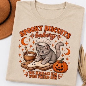 Può includere: Una t-shirt beige con la grafica di un gattino grigio con un cappello da chef che impasta su un tagliere di legno. Il testo "SPOOKY BISCUITS bakery" e "WE KNEAD EM YOU NEED EM" è in arancione, accompagnato da una zucca, stelle e una luna crescente.