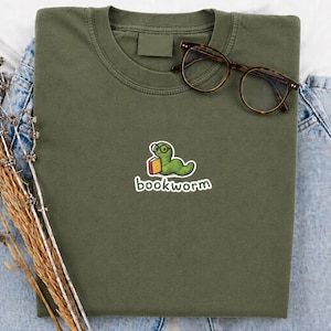Può includere: Una t-shirt verde oliva con il ricamo di un verme verde che indossa occhiali tondi e tiene in mano un libro rosso e giallo. Sotto il verme, la parola bookworm è ricamata in bianco. Un paio di occhiali marroni è appoggiato sulla t-shirt.