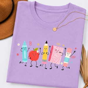Può includere: Una t-shirt viola chiaro con una stampa grafica di cinque articoli scolastici antropomorfi, tra cui un pastello blu, una mela rossa, una matita gialla, un quaderno rosa e una colla stick, tutti con volti sorridenti.