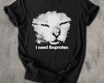 May include: A black short-sleeved t-shirt featuring a white, distressed-style graphic of a lion's face in the center. Below the lion graphic, the text "I need ibuprofen" is printed in a clean, white, sans-serif font on the chest area.