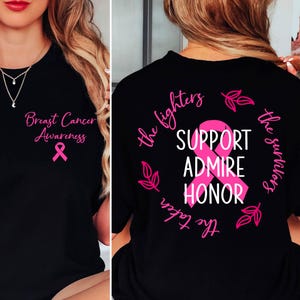 Può includere: T-shirt nera con testo e nastro rosa per la sensibilizzazione sul cancro al seno. La parte anteriore della maglietta recita "Breast Cancer Awareness" con un nastro rosa. La parte posteriore della maglietta ha un nastro rosa e le parole "Support, Admire, Honor".