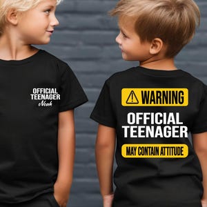 May include: Two black t-shirts with white text. One shirt says "OFFICIAL TEENAGER Noah" on the front. The other shirt has a yellow warning sign with the text "WARNING OFFICIAL TEENAGER MAY CONTAIN ATTITUDE" on the back.