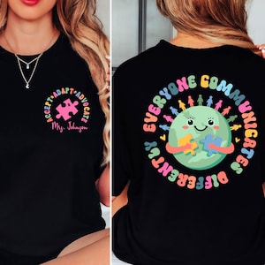 Puede incluir: Camiseta negra con un diseño colorido. La parte delantera presenta una pieza de rompecabezas con las palabras "ACCEPT • ADAPT • ADVOCATE" y el nombre "Mrs. Johnson". La parte trasera muestra una Tierra sonriente sosteniendo piezas de rompecabezas, rodeada por el texto "EVERYONE COMMUNICATES DIFFERENTLY".