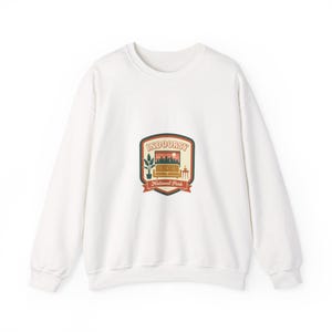 Peut inclure: Sweat-shirt blanc avec un motif graphique sur le devant. Le motif est un écusson avec le texte "INDOORSY National Park". Le motif représente un canapé, une plante, une table et un paysage urbain avec un coucher de soleil.