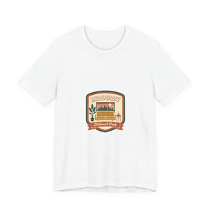 May include: White t-shirt with a graphic that reads "INDOORSY National Park." The design shows a living room scene with a sofa, plant, and side table. The graphic uses brown, orange, and green colours.