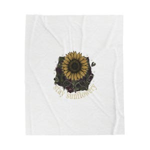 Pode incluir: Cobertor branco com um desenho de girassol e o texto "stay sunflowery". O girassol é amarelo e castanho, sobre um fundo verde escuro e bordô.