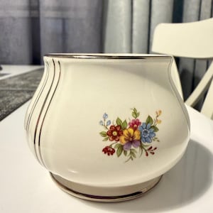 May include: Cream-colored ceramic bowl with a floral design. The bowl has a rounded shape with a wide base and a slightly flared rim. The floral design features red, yellow, blue, and purple flowers. Gold trim accents the rim and vertical lines.