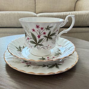May include: A white teacup, saucer, and dessert plate set. The set features a floral design with pink flowers and green leaves. The edges of the plates and cup are trimmed with gold. The teacup has a curved handle.