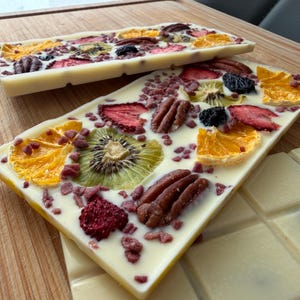 Op de afbeelding: Twee rechthoekige repen witte chocolade belegd met gedroogd fruit en noten. De toppings bestaan uit schijfjes gedroogde sinaasappel, kiwi, aardbeien, bramen en hele pecannoten, bestrooid met kleine stukjes rode gedroogde vruchten.