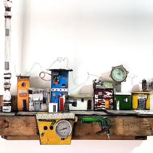 May include: A mixed-media artwork depicting a miniature townscape with vibrant buildings, wiring, and a wooden base. The piece includes a tall, white and brown vertical element and a yellow gauge. The artist's signature is visible.