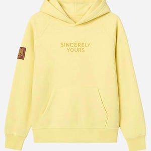 May include: A pale yellow hoodie with a front pocket and a hood. The words "SINCERELY YOURS" are embroidered in gold thread across the chest. A small patch is on the left sleeve. This casual sweatshirt is perfect for everyday wear.