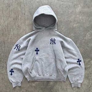May include: A heather gray hooded sweatshirt featuring dark navy blue embroidered accents. The design includes the New York Yankees logo on the chest and right sleeve, along with dark navy blue cross symbols on the chest and both sleeves.