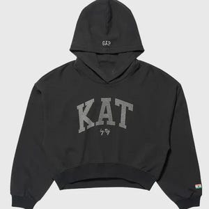 Puede incluir: Una sudadera negra corta con capucha que tiene el texto GAP bordado en blanco. El frente muestra la palabra KAT en pedrería plateada con el texto by Raj debajo. Un pequeño parche rectangular con una bandera es visible en la manga izquierda.