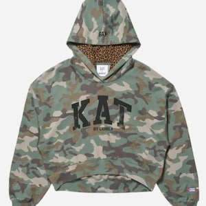 Può includere: Una felpa corta con cappuccio caratterizzata da un motivo mimetico verde, marrone e beige. La parte anteriore mostra la scritta nera KAT BY DANIELA. L'interno del cappuccio ha una stampa leopardata marrone e la parola GAP è ricamata sul cappuccio. Una piccola bandiera è presente sulla manica.