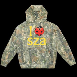 May include: A camouflage print hooded sweatshirt featuring a large yellow graphic that reads I SZA with a red ladybug with black spots replacing the heart symbol. The hoodie has long sleeves, a front kangaroo pocket, and a drawstring hood.