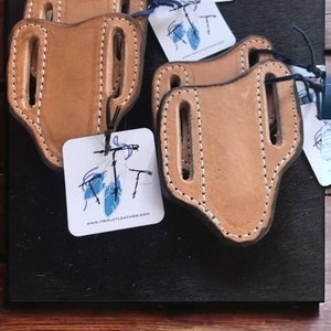 May include: Four brown leather knife sheaths with stitching detail. The sheaths are stacked on top of each other. A white tag with blue feathers and the text "www.tripletleather.com" is visible in the bottom right corner of the image.