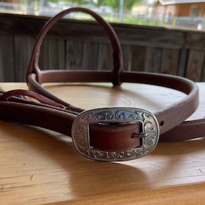 May include: A brown leather horse halter with a silver buckle featuring a decorative floral design.