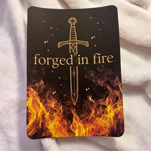 May include: A rectangular card with a black background featuring a golden sword design and the words "forged in fire." The bottom of the card displays flames in shades of orange and yellow, with small white stars scattered across the top.