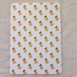May include: A rectangular white item with rounded corners, covered in a repeating pattern of orange pineapples with blue tops. The pineapples are evenly spaced across the surface, creating a tropical and playful design.