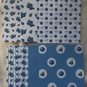 May include: A collection of patterned cards featuring blueberry designs. The cards display various patterns, including blueberries on white, bows and dots, stripes, and a blue background with blueberry motifs. The color scheme is primarily blue and white.