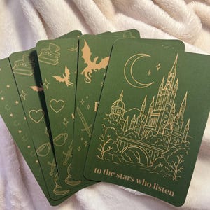 May include: A collection of green cards with gold illustrations of dragons, castles, and other fantasy-themed images. The cards have a crescent moon, stars, and the text "to the stars who listen". The cards are arranged on a soft, white blanket.