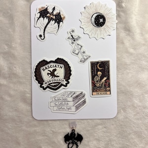 May include: A collection of stickers and a charm featuring fantasy and celestial themes. Stickers include a dragon, a sun and moon design, a sword, a crest that says "Basgiath War College", a tarot card, and a stack of books. A black dragon charm with a star pendant is at the bottom.