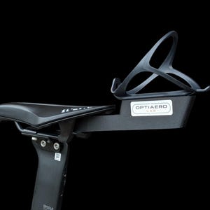 Op de afbeelding: Een zwart fietszadel gemonteerd op een zadelpen met een bidonhouder aan de achterzijde. De montagebeugel heeft een wit rechthoekig label met de tekst OPTIAERO LAB in zwarte letters. Het zadel is voorzien van de merknaam fi'zi:k.