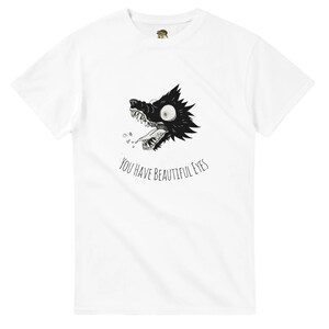 Può includere: T-shirt bianca con un'illustrazione in bianco e nero della testa di un lupo con la bocca aperta e la scritta "YOU HAVE BEAUTIFUL EYES". Il lupo ha grandi occhi e denti affilati.