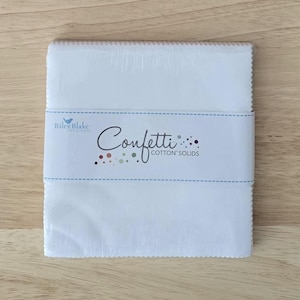May include: A stack of white fabric squares wrapped in clear plastic with a paper band. The band features the Riley Blake Designs logo and the text Confetti Cotton Solids with small colorful dots. The fabric edges have a pinked, zigzag finish.