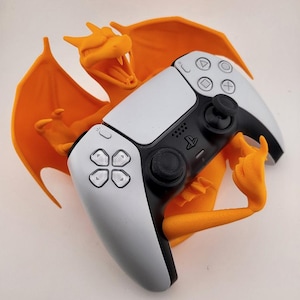Könnte beinhalten: Eine orangefarbene, 3D-gedruckte Charizard-Figur, die einen weiß-schwarzen PlayStation 5-Controller hält. Der Controller hat einen schwarzen Joystick, Tasten und ein PlayStation-Logo. Das Charizard hat ausgebreitete Flügel und einen offenen Mund.