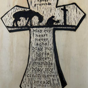 May include: Wooden cross with a black outline and etched design. The cross features a horse, a person kneeling, and a cross silhouette. Text includes "May my belly never grumble" and "Amen."