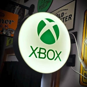 May include: A circular, wall-mounted, illuminated sign featuring the green Xbox logo and the word XBOX in green text on a white, glowing background. The sign has a black outer frame and is connected to a white power cord extending to the right.