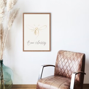 Könnte beinhalten: Ein gerahmter Druck mit einer goldfarbenen Bienenillustration und dem Text "Bee classy". Das Kunstwerk befindet sich auf einem cremefarbenen Hintergrund und ist in einem hellbraunen Holzrahmen gerahmt. Der Druck wird an einer weißen Wand angezeigt. Dazu ein brauner Ledersessel.